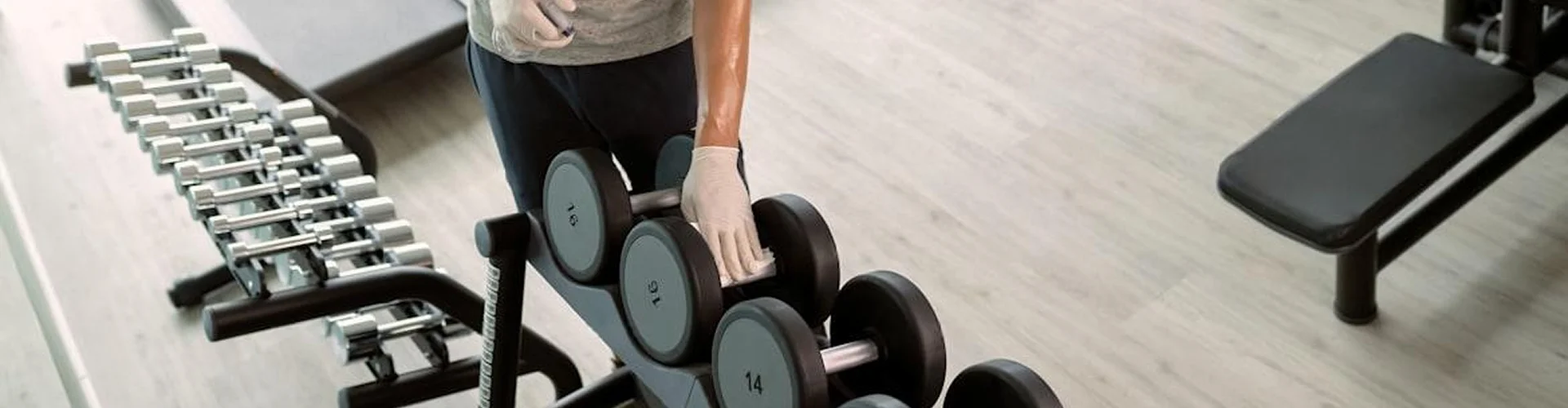 A gloved hand wiping down a rubber dumbbell rack beside a chrome dumbbell tree and a bench