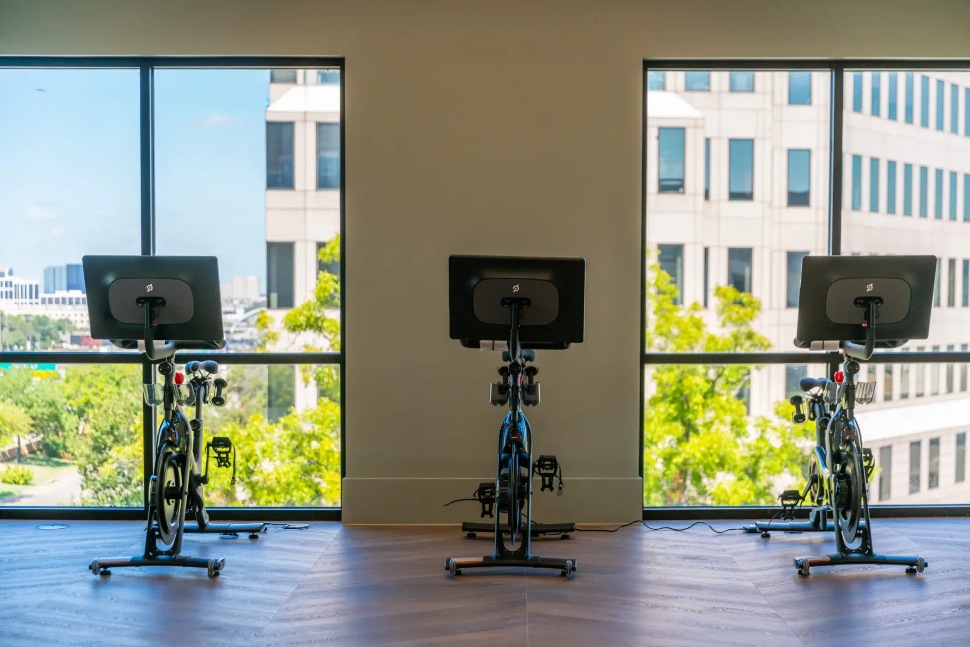 Indoor cycling bikes with screens against a window wall in a Texas apartment gym, on wood-look flooring