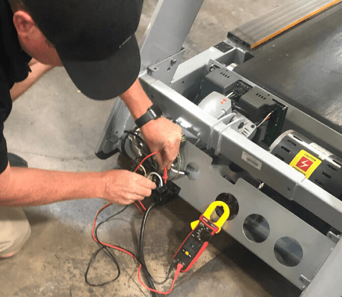 Technician testing a treadmill motor with a clamp meter, the motor and controller exposed under the deck