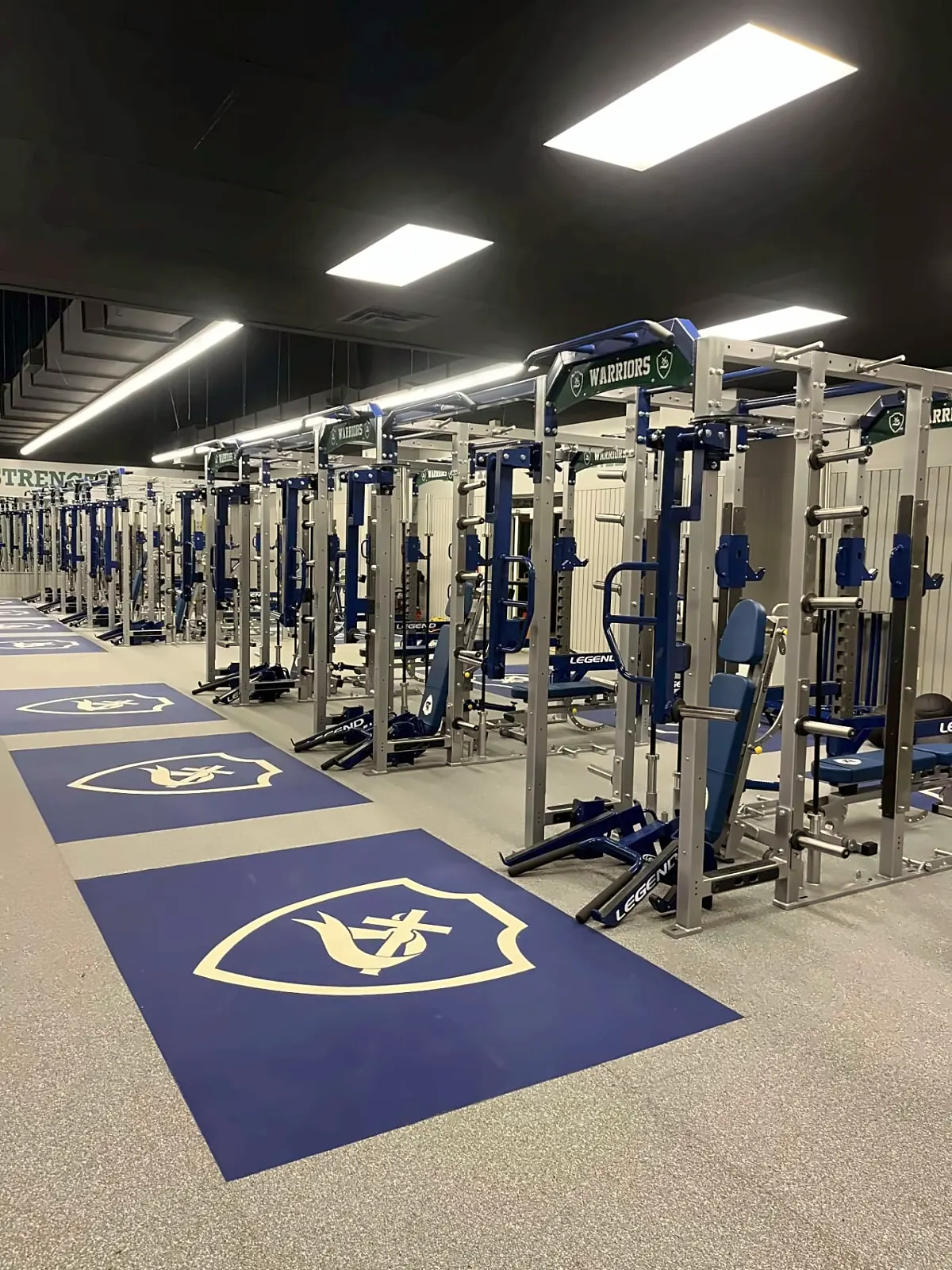 Little Rock Christian Academy weight room: rows of silver and blue racks behind navy shield-logo platforms