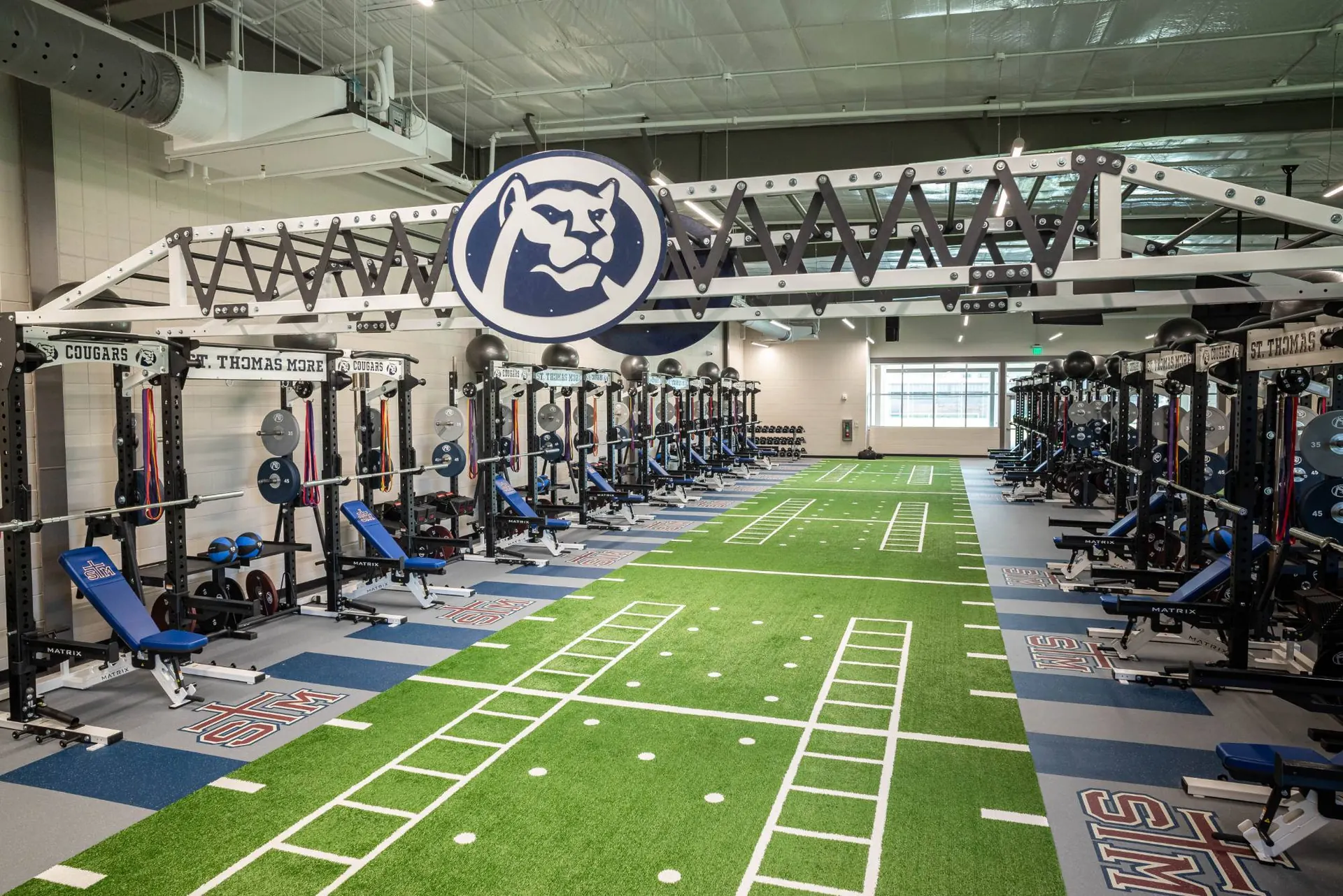 St. Thomas More weight room in Lafayette, LA: facing rack rows and a turf lane under a truss with the Cougars emblem