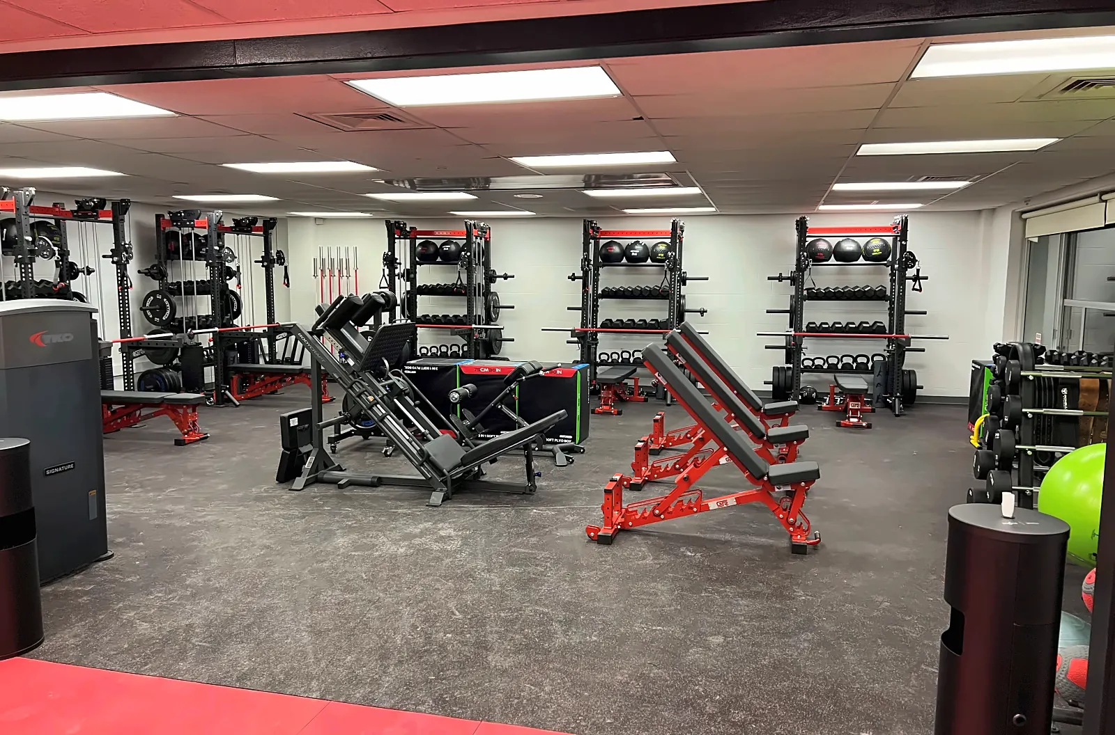 Saranac High School weight room: a hack squat and red incline benches in front of a row of storage racks
