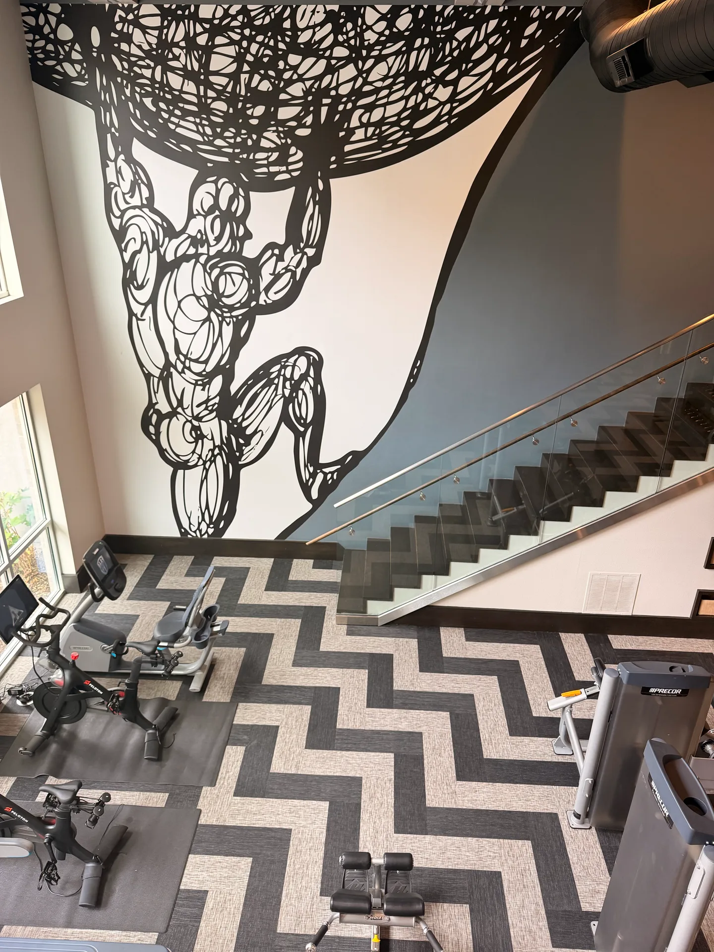 Chevron plank floor seen from the mezzanine, with indoor cycles below a figure mural in a Dallas apartment gym