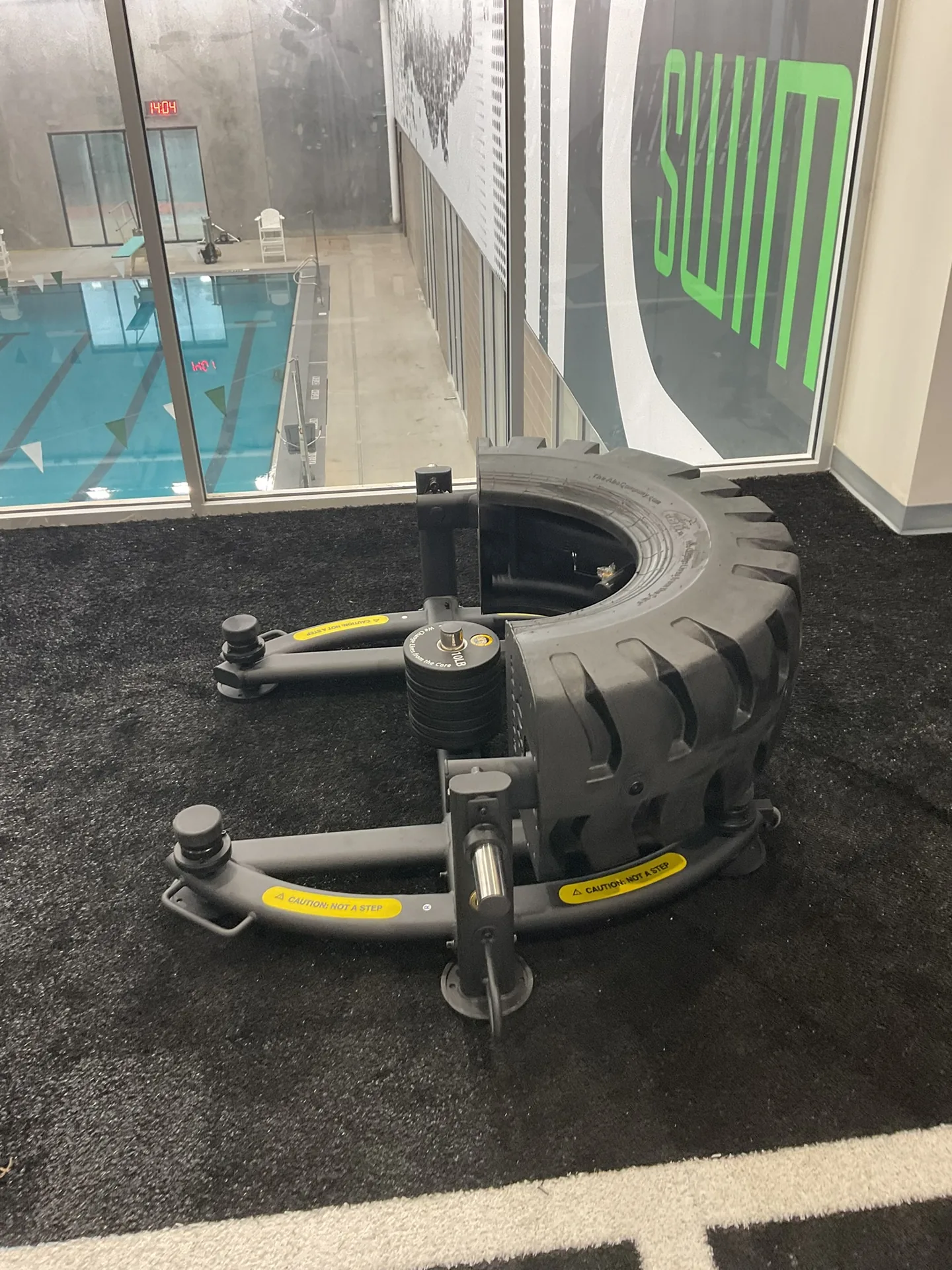 A tire-flip trainer on black turf beside a glass wall overlooking a competition swimming pool