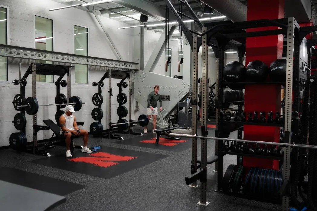 Rolled rubber flooring and lifting platforms across a college weight room with racks, bumper plates and dumbbells