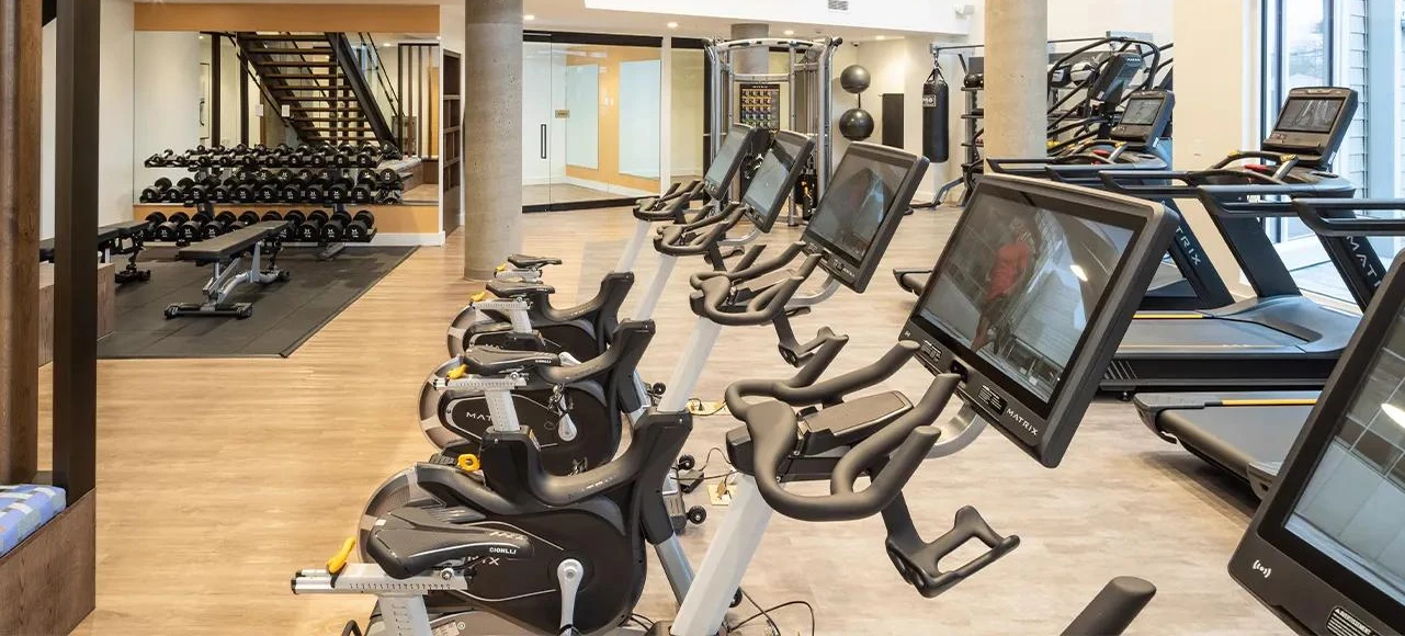 Apartment gym floor with a row of Matrix bikes and treadmills, a dumbbell rack and a rig with a hanging heavy bag