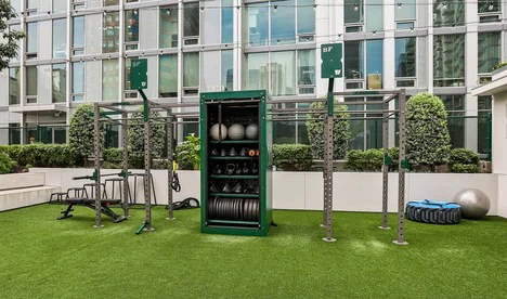 Courtyard outdoor gym on turf with a multi-station rig, storage cabinet and medicine balls beside an apartment building