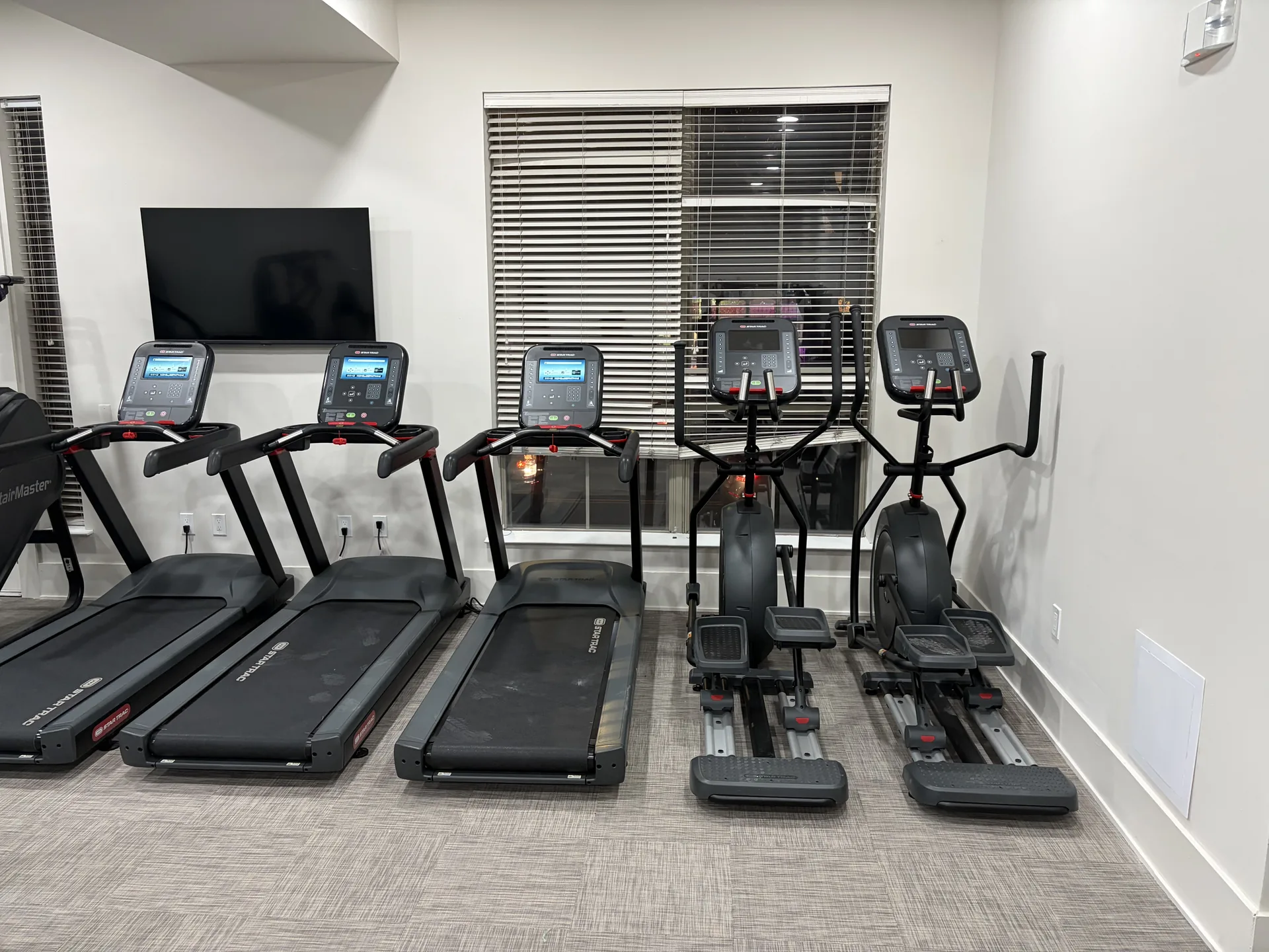Cardio row of treadmills, an elliptical and a cross trainer beneath a wall-mounted screen at a Philadelphia apartment gym