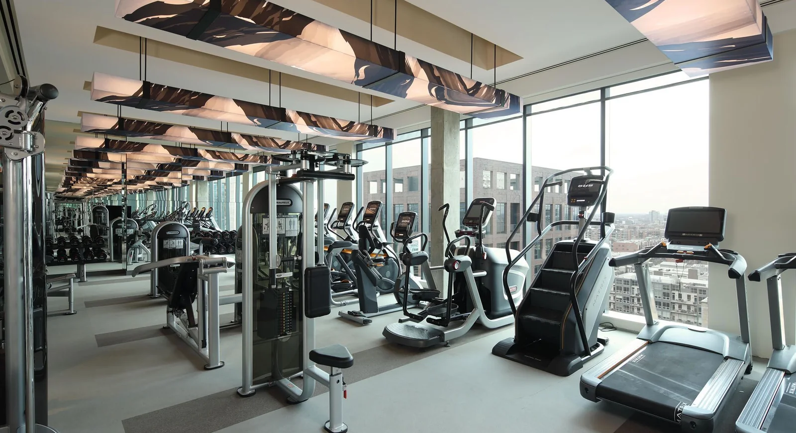 Rows of treadmills, ellipticals and stair climbers on a large fitness floor beside a floor-to-ceiling glass wall