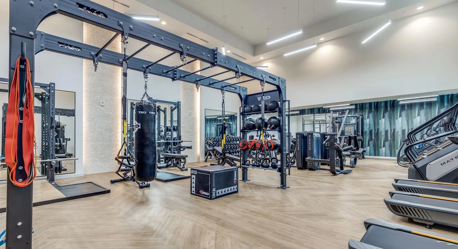 Fitness floor with a TKO functional rig, heavy bags, dumbbell racks and Matrix cardio over wood-look plank