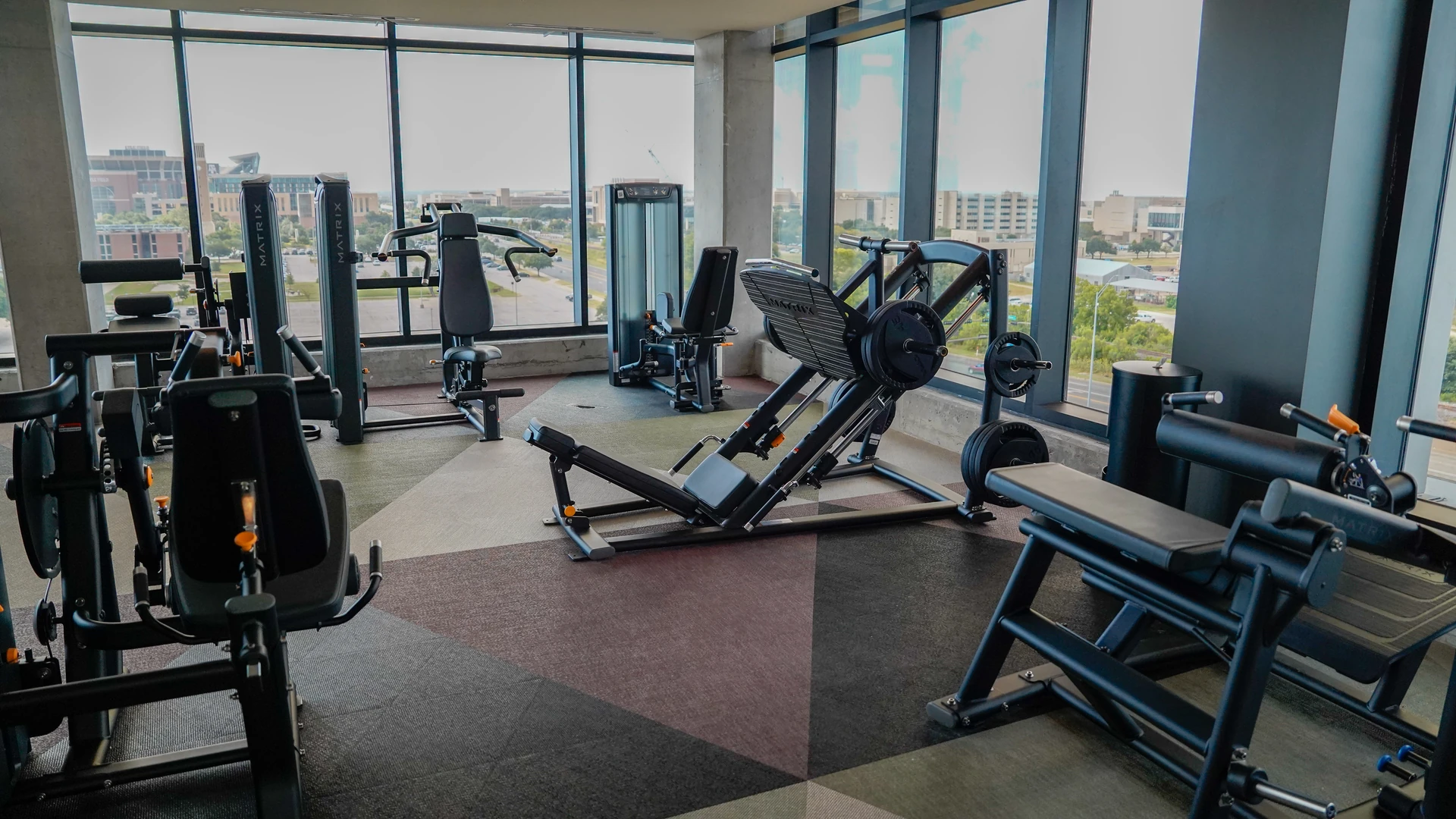 Matrix leg press and plate-loaded machines facing floor-to-ceiling windows in a student housing gym