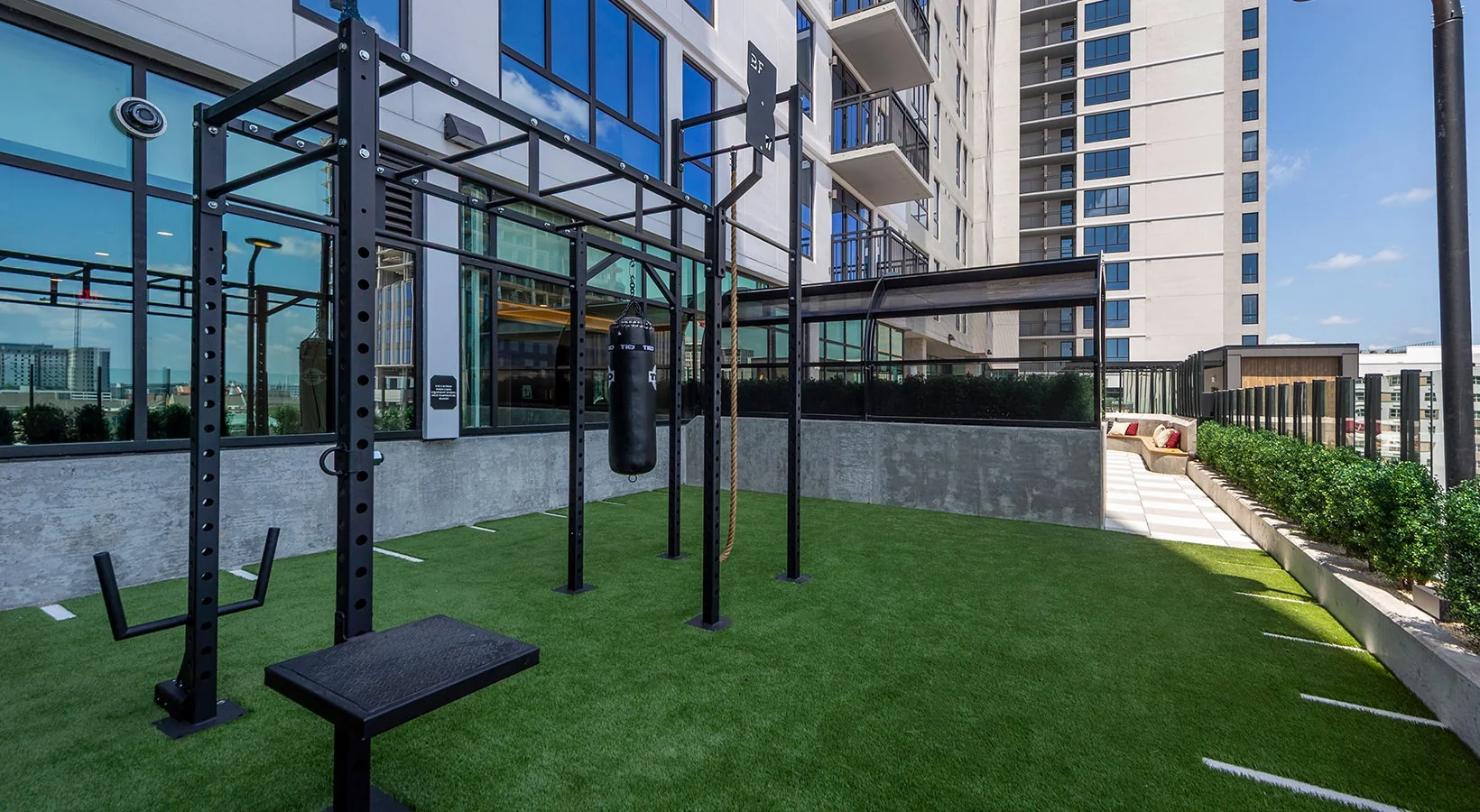Outdoor rooftop training rig on turf with a rope climb and heavy bag at a student housing wellness deck